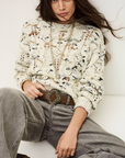 A woman with long brown hair wears the BA&SH Jumper Rave, a cream-colored, patterned knit sweater, paired with light gray jeans, a brown ornate-buckle belt, and layered necklaces. She sits on a white surface, looking forward neutrally.