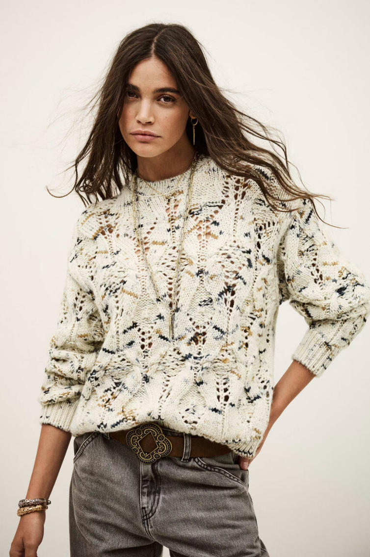 A young woman with long brown hair wears the BA&SH Jumper Rave, a cream and blue patterned 100% wool sweater, styled with grey jeans, a decorative belt, and gold bracelets, posing with one hand on her hip against a plain background.
