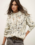 A young woman with long brown hair wears the BA&SH Jumper Rave, a cream and blue patterned 100% wool sweater, styled with grey jeans, a decorative belt, and gold bracelets, posing with one hand on her hip against a plain background.