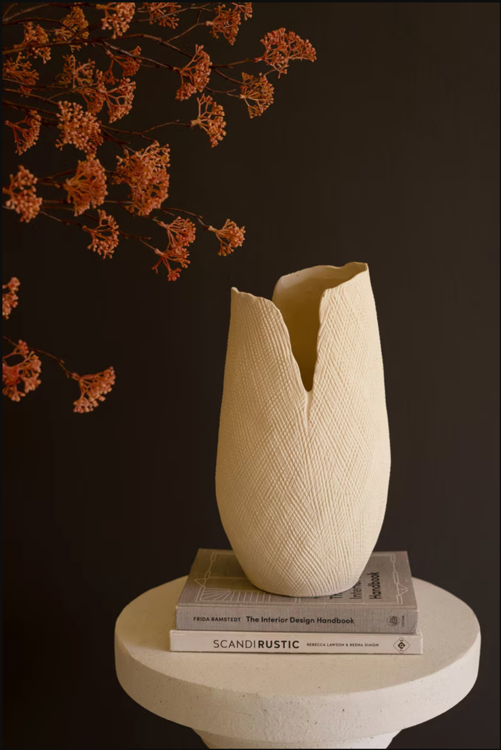 The Bloom Flower Vase L by Kalalou, Inc—a beige, textured vase with an irregular opening—rests on two books atop a round white pedestal, displaying rust-colored dried flowers against a dark brown backdrop.