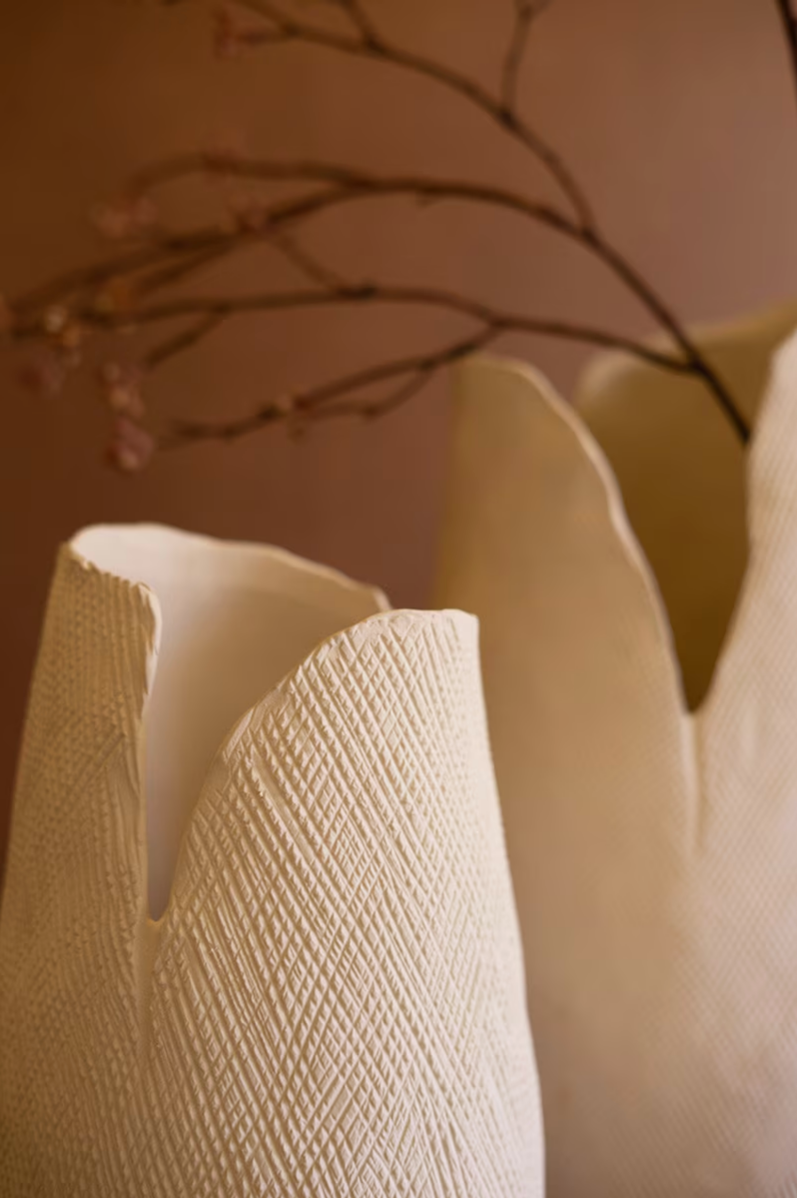 The Bloom Flower Vase S by Kalalou, Inc features a white textured finish with jagged, organic edges. Against a soft brown background, it holds thin branches with small dried flowers, adding a natural, minimalist charm to any space.