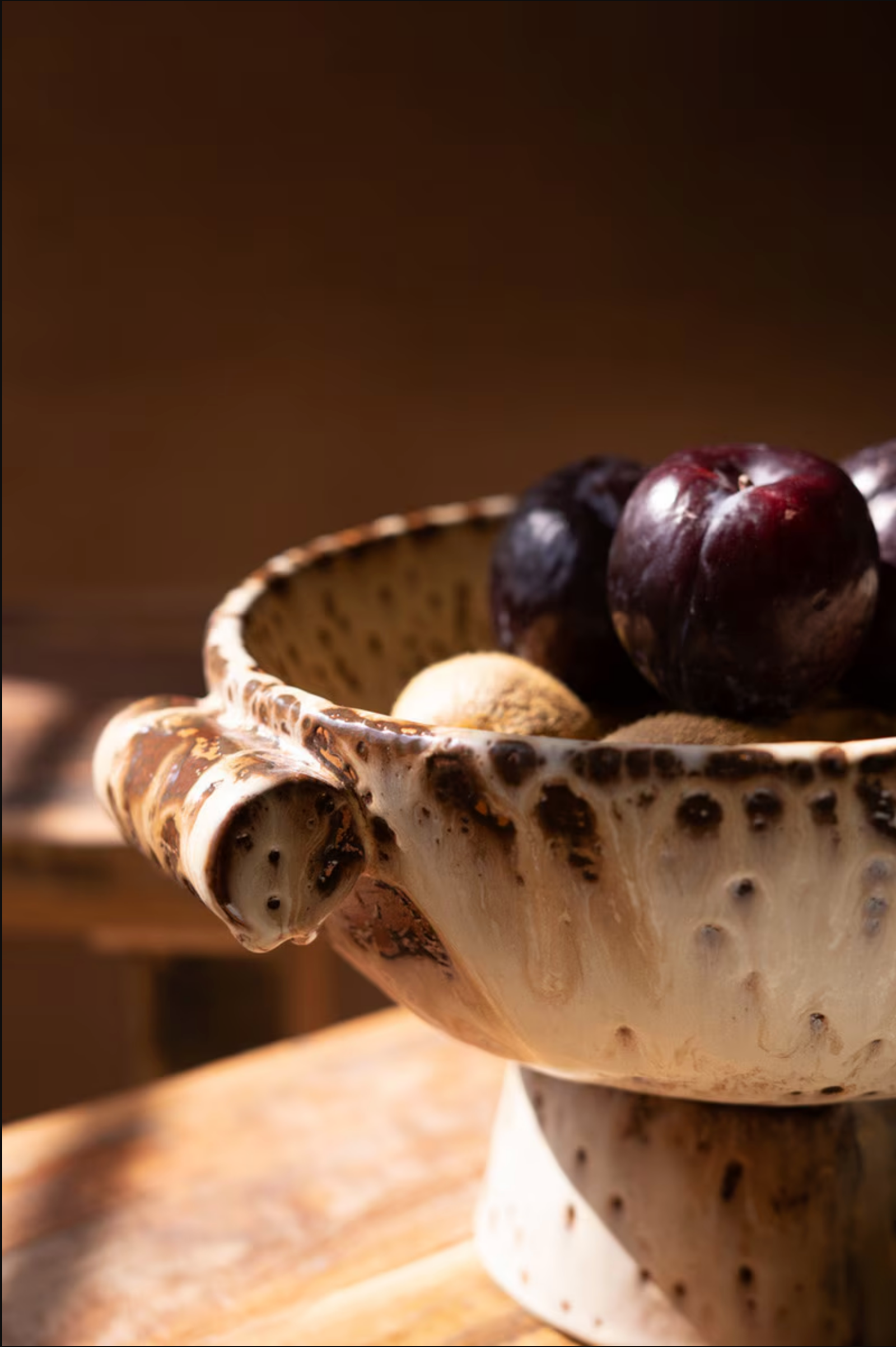 The Kalalou, Inc Rustic Ceramic Compote, filled with dark purple plums and yellow-brown fruits, rests on a wooden surface in warm light. This kitchen decor piece stands out in focus against a softly blurred background.