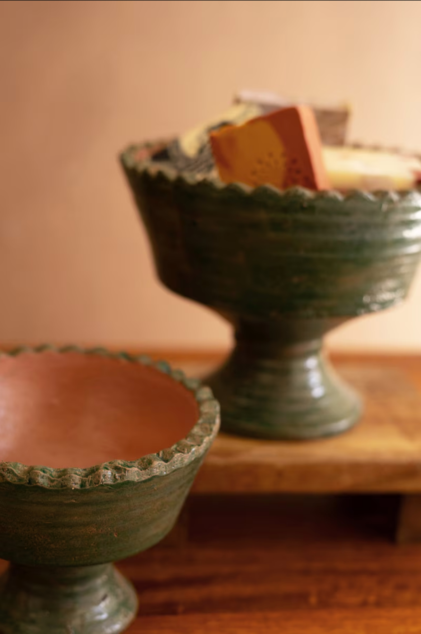 Two Green Terra Clay Compote bowls by Kalalou, Inc sit on wood; one holds colorful cheese blocks, the other is empty. A blurred, warm-toned background enhances the inviting decorative scene.