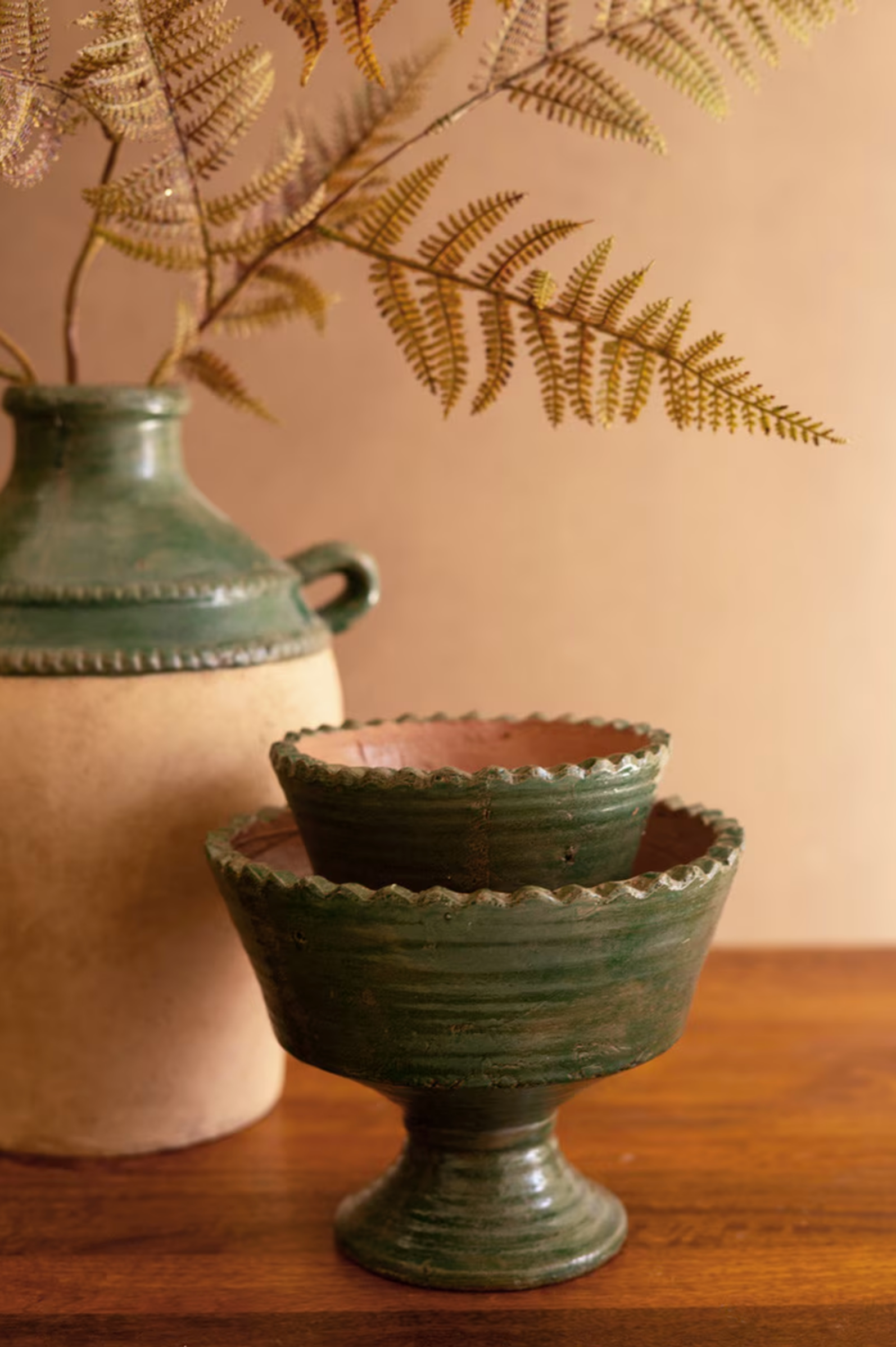 The Green Terra Clay Compote by Kalalou, Inc is stacked on a wooden surface, paired with a beige and green vase holding dried ferns for a rustic, earthy still-life display.