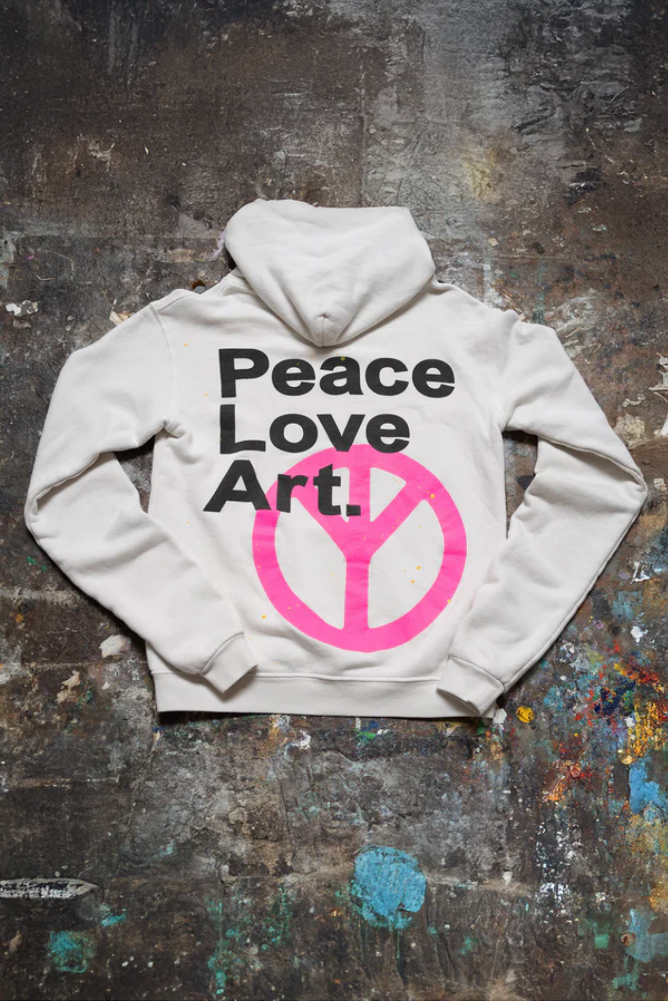 The Made Worn Peace Love Oversized Hoodie rests on a paint-splattered surface. Its back features bold black "Peace Love Art." text above a large pink peace symbol, making it a cozy winter pick for creative spirits.