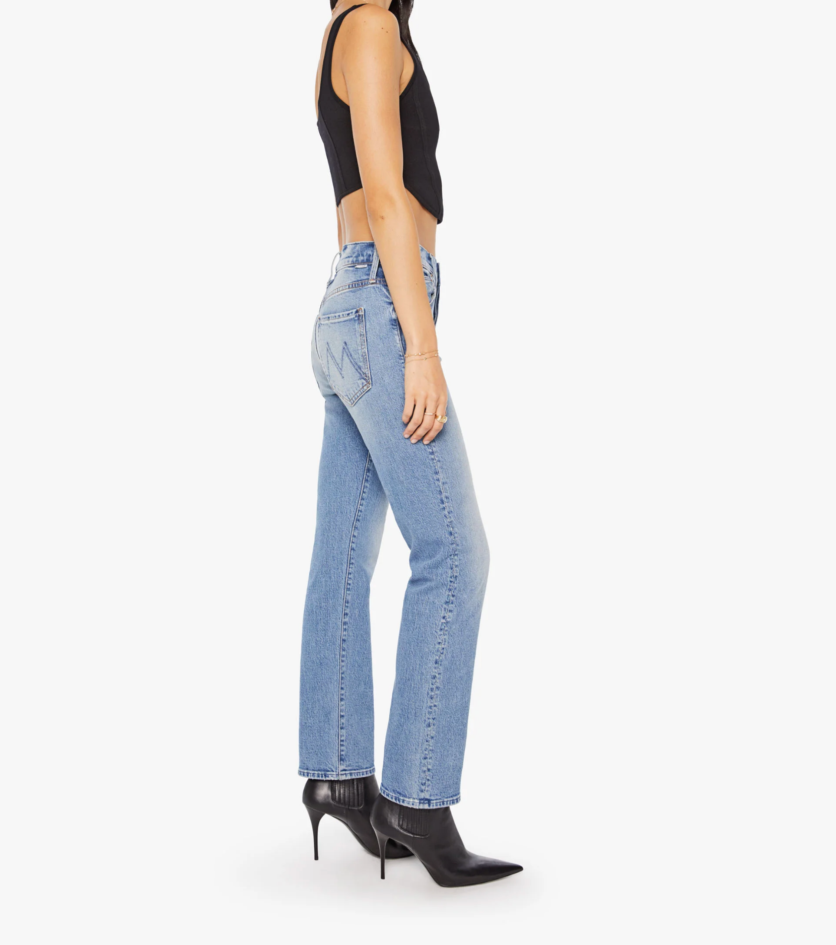 A person wearing a sleeveless black top, black pointed-toe heeled boots, and Mother Mid Rise Hiker Hover jeans featuring a zigzag design on the back pocket, stands sideways against a white background.