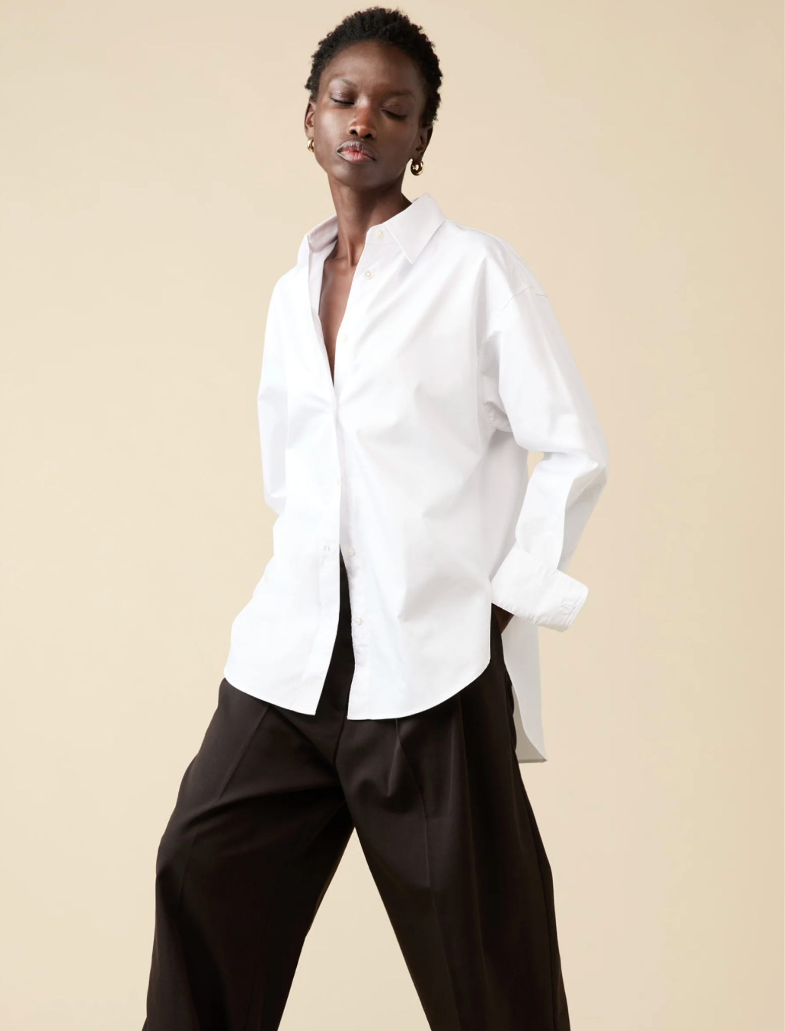 Against a beige background, a person wears Sold Out NYC’s oversized white The Everything Shirt and wide-leg black pants, eyes closed with one hand in a pocket.