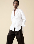 Against a beige background, a person wears Sold Out NYC’s oversized white The Everything Shirt and wide-leg black pants, eyes closed with one hand in a pocket.