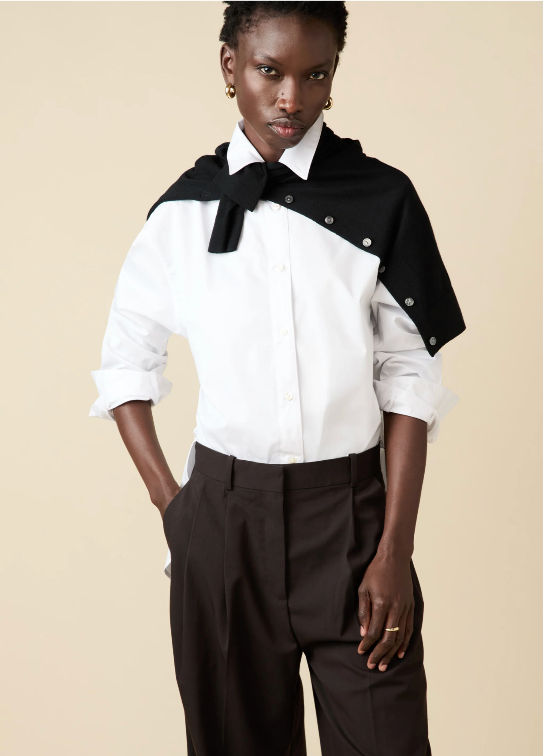A person stands with one hand in their pocket, wearing Sold Out NYC’s The Everything Shirt— a tailored 100% cotton white button-up—paired with dark brown trousers and a black sweater draped over their shoulders against a beige background.