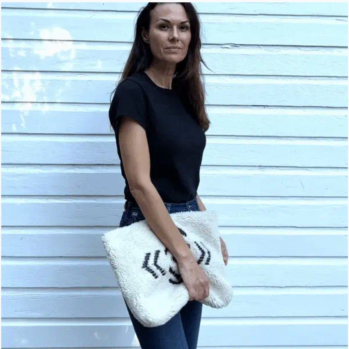 A woman in a black t-shirt and blue jeans stands before a white wooden wall, holding the Kempton & Co. Oversized Clutch featuring a black spider design.