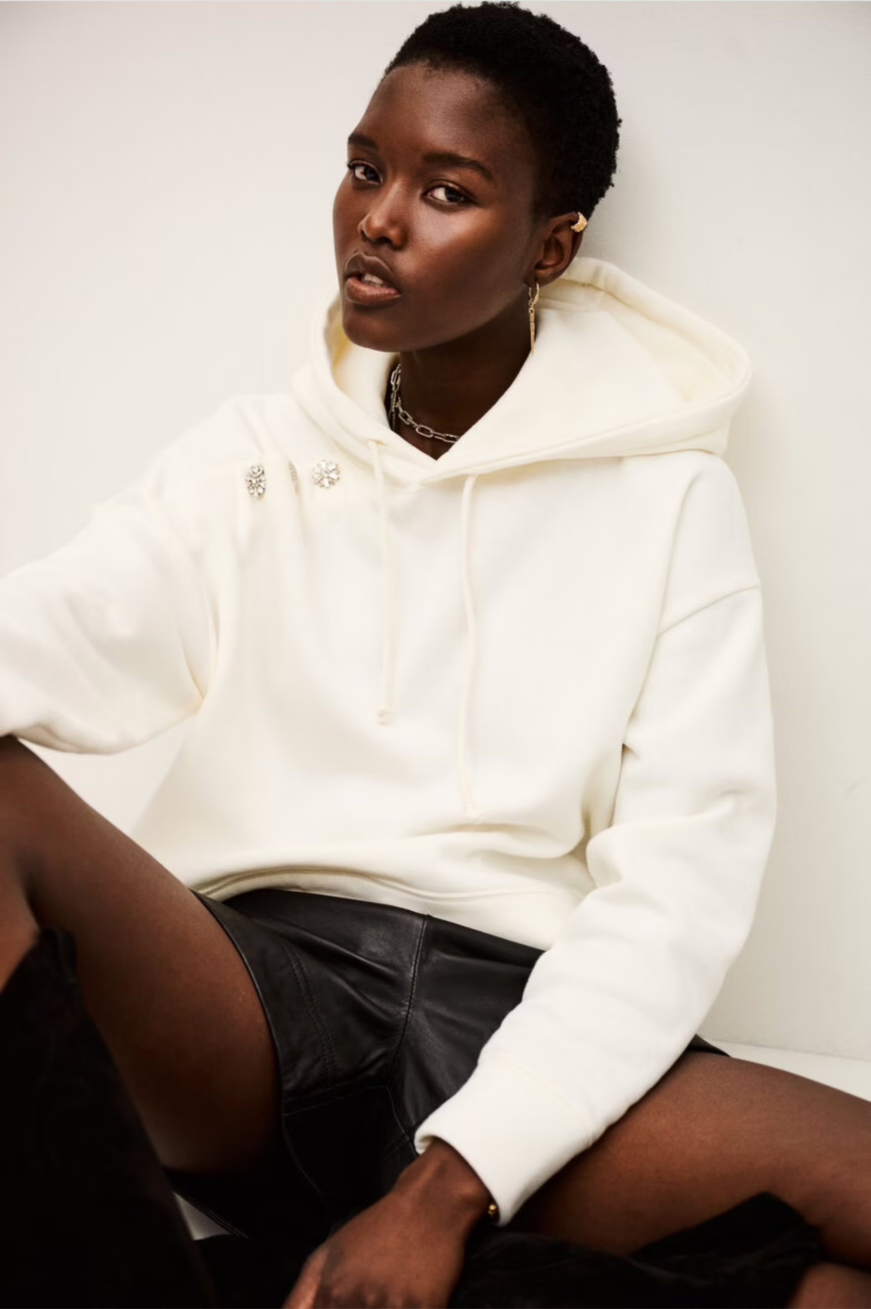 A person with short hair wears the BA&SH Sweatshirt Didi—a white, washable polyester cotton hoodie—styled with pins, black leather shorts, and a chain necklace, sitting against a white background.