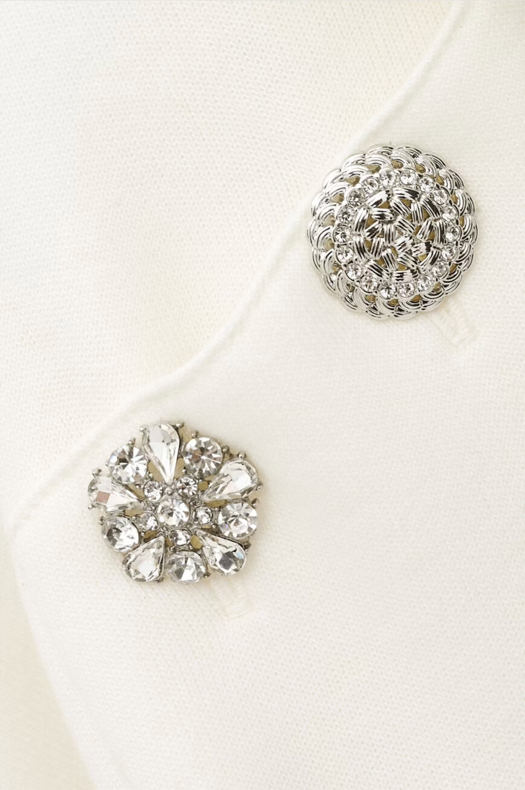 Close-up of two decorative silver buttons on the BA&SH Sweatshirt Didi: one with a woven circular pattern, the other embellished with clear faceted gemstones in a floral design, set against white polyester-cotton fabric.