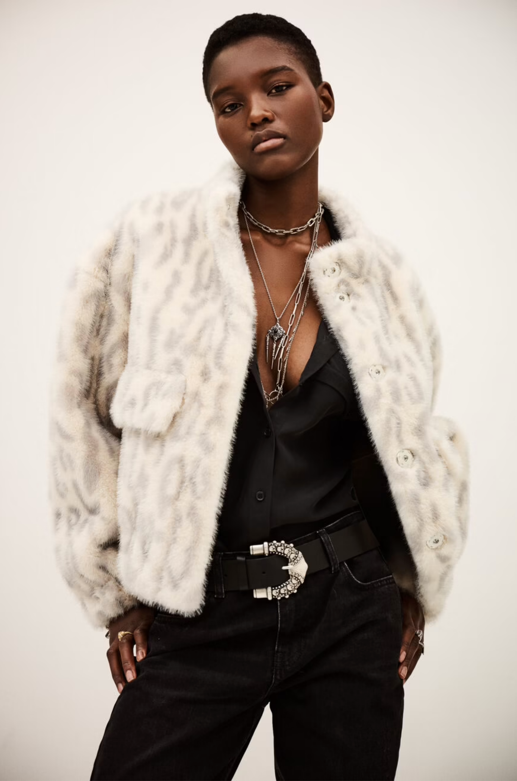 Wearing the BA&SH Coat Saros, a light leopard-print faux fur cropped jacket, over a low-cut black top with silver necklaces, a stylish belt, and black jeans, a person poses confidently against a plain backdrop.