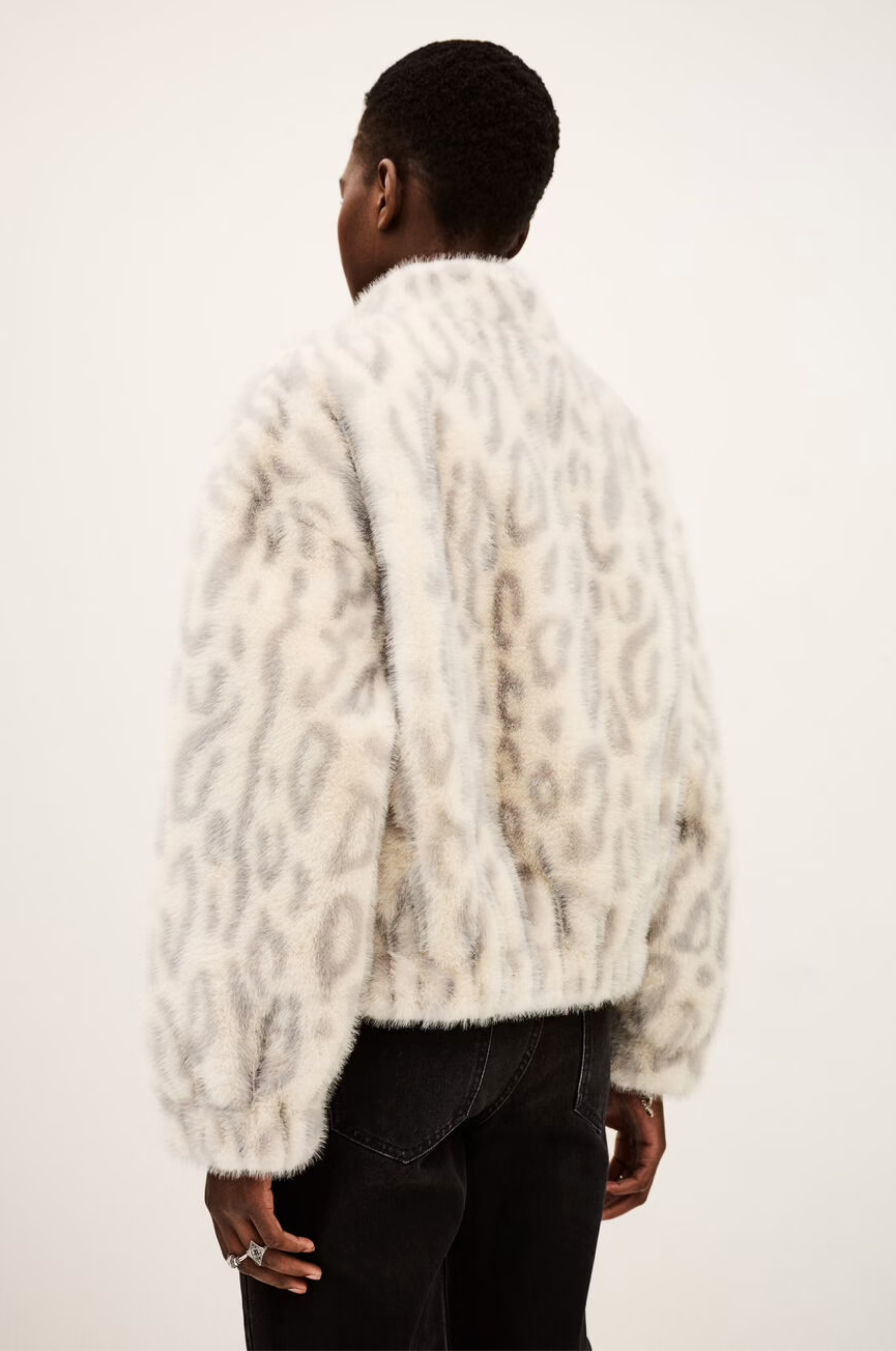 Someone stands facing away, wearing the BA&SH Coat Saros—a printed faux fur cropped jacket with grey animal prints—paired with black jeans against a plain light background.