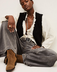 A person sits cross-legged on the floor, wearing BA&SH Jean Yusc wide-leg gray jeans with a white cotton ruffled blouse, black knit vest, belt, brown boots, and silver jewelry against a plain light background.