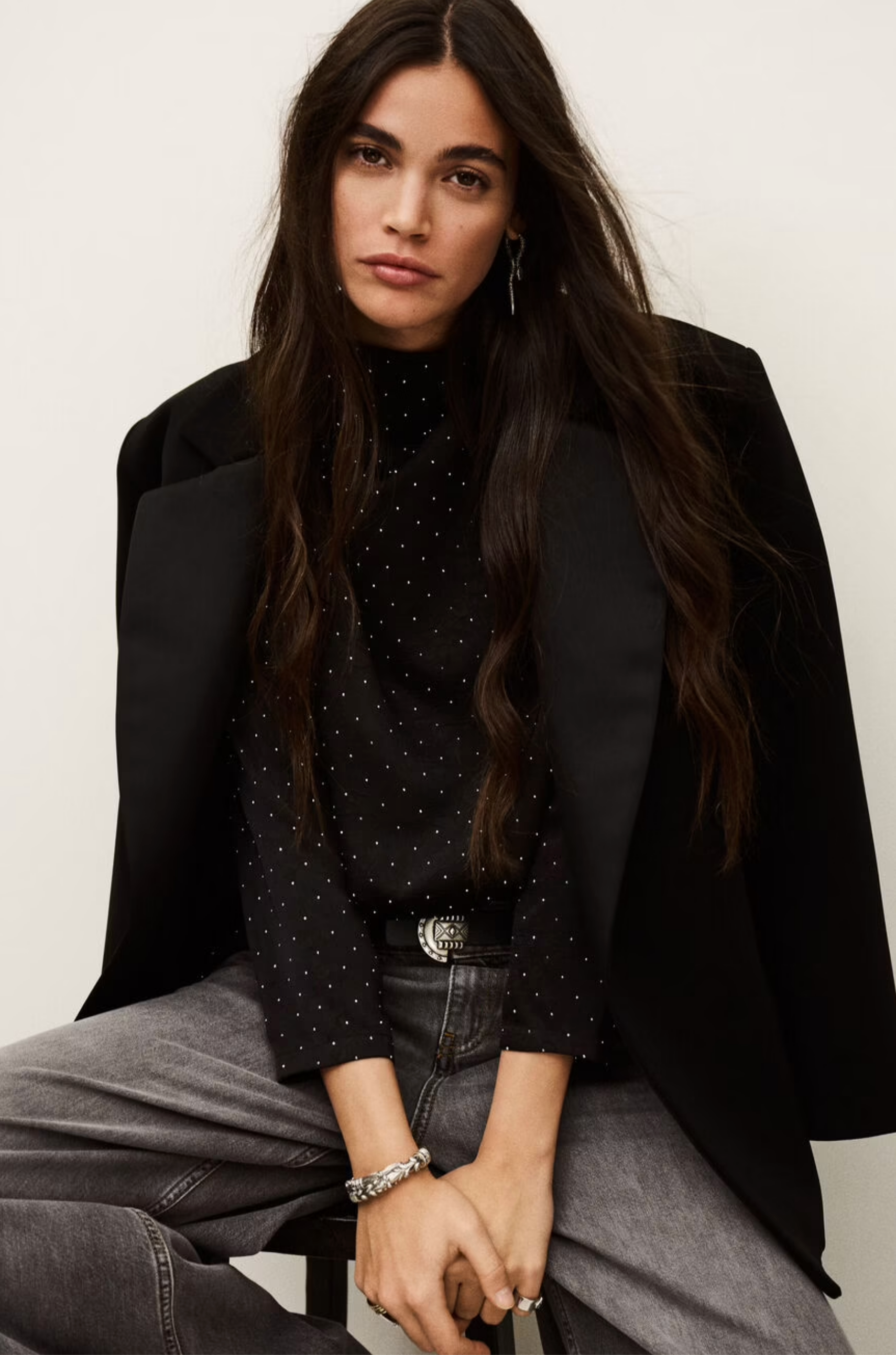 A woman with long dark hair sits on a stool, wearing the BA&SH Top Debra—a black top with mini studs—paired with a black batwing-sleeve blazer, gray jeans, and silver jewelry. She looks directly at the camera with a neutral expression.