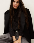 A woman with long dark hair sits on a stool, wearing the BA&SH Top Debra—a black top with mini studs—paired with a black batwing-sleeve blazer, gray jeans, and silver jewelry. She looks directly at the camera with a neutral expression.