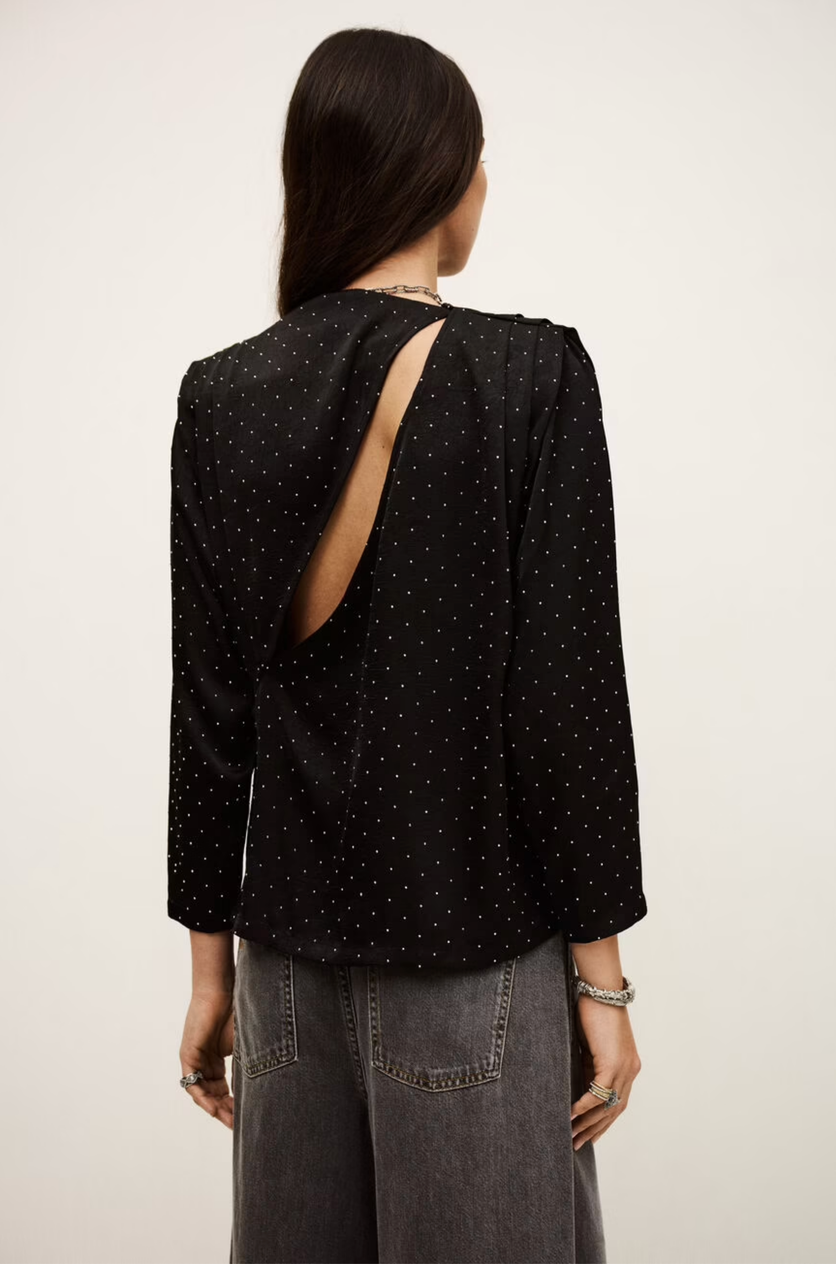 A person with long dark hair is seen from behind, wearing the BA&SH Top Debra—a black, long-sleeve batwing blouse with mini studs and an asymmetrical open back—paired with high-waisted, loose gray jeans.