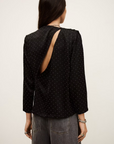 A person with long dark hair is seen from behind, wearing the BA&SH Top Debra—a black, long-sleeve batwing blouse with mini studs and an asymmetrical open back—paired with high-waisted, loose gray jeans.
