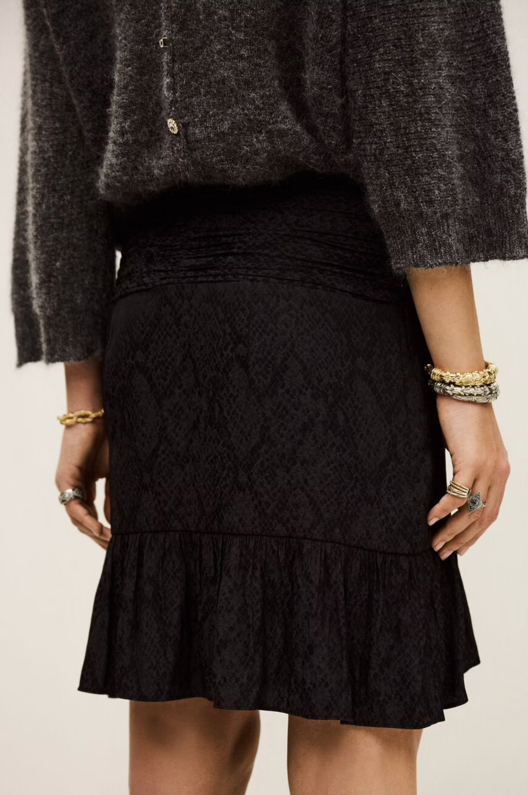 A person wears a textured dark grey polyester sweater and the BA&SH Skirt Rino—black, patterned with a ruffled hem. Gold bracelets and rings adorn their wrists and fingers. Image is shoulder to knee; dry clean recommended.