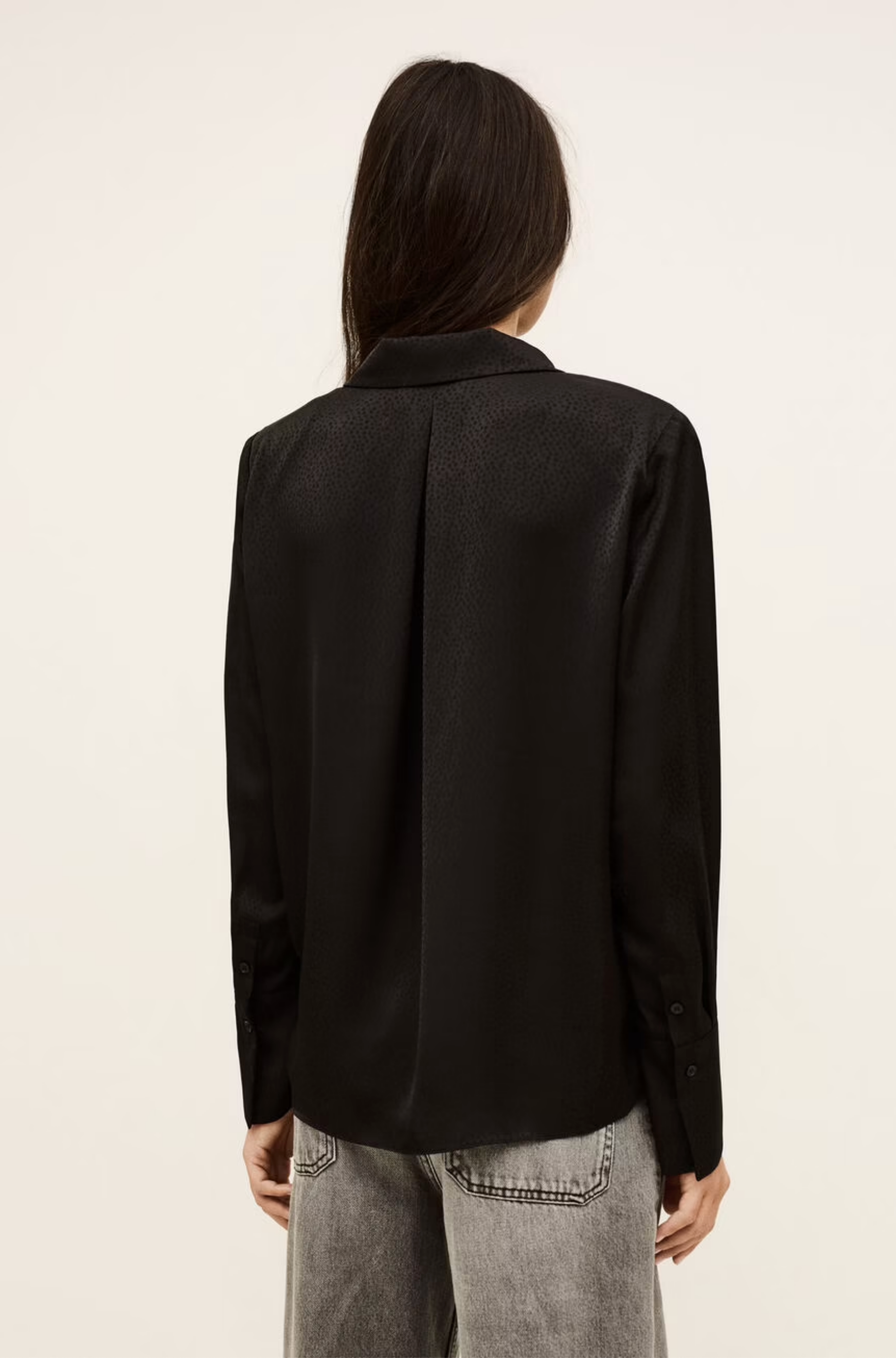 A person with long dark hair is shown from the back against a plain light background, wearing the BA&SH Shirt Itsey—a black long-sleeve V-neck top—paired with light grey jeans.