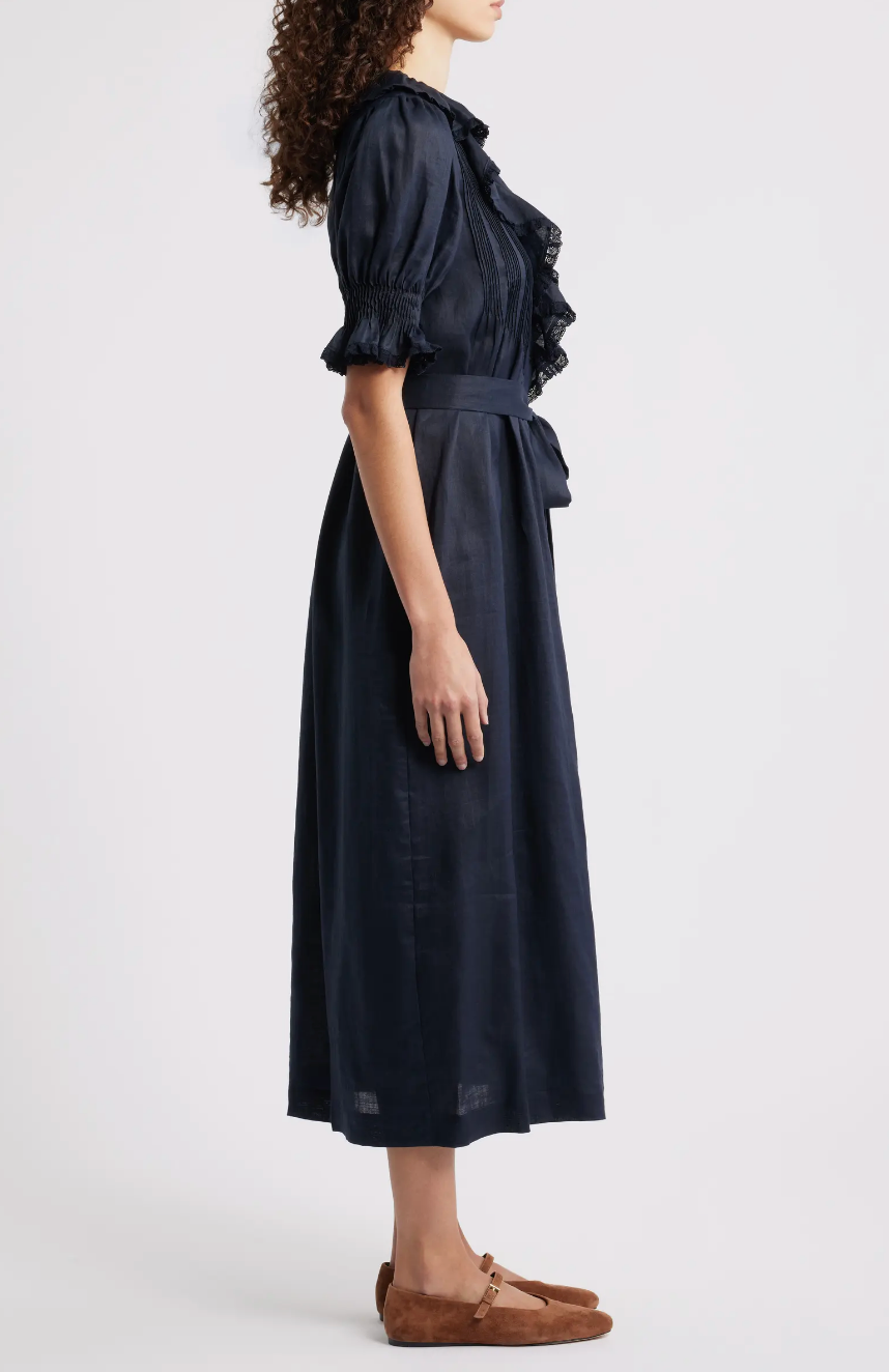 A woman stands in profile against a plain white background, wearing the DOEN Harriette Dress in navy with ruffled sleeves and a belted waist, styled with brown flats. Her curly hair enhances the elegant look.