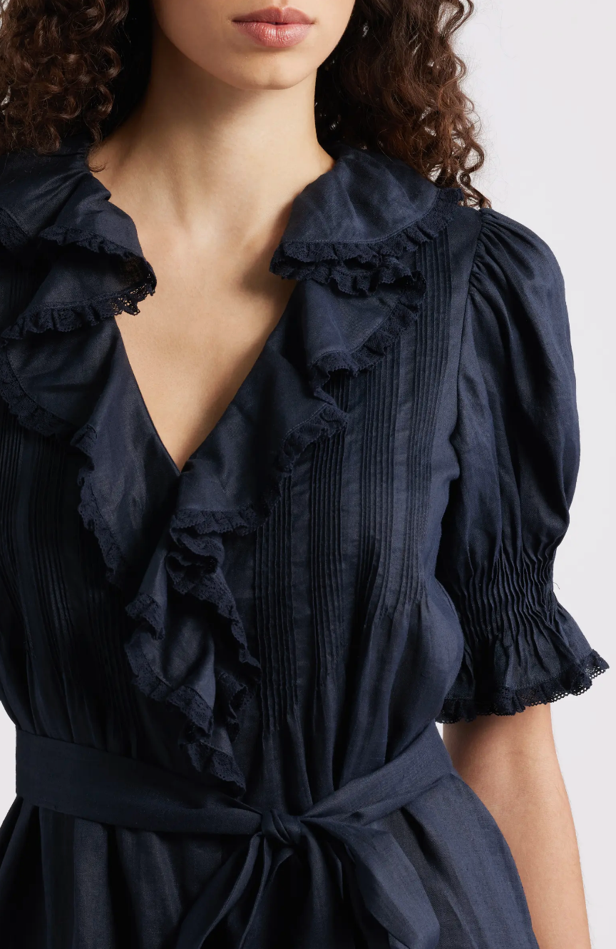 A woman models the DOEN Harriette Dress, featuring navy blue fabric, ruffled collar and front, puffed short sleeves with shirred cuffs, and a waist-cinching belt. Her curly hair frames her shoulders for an elegant look.