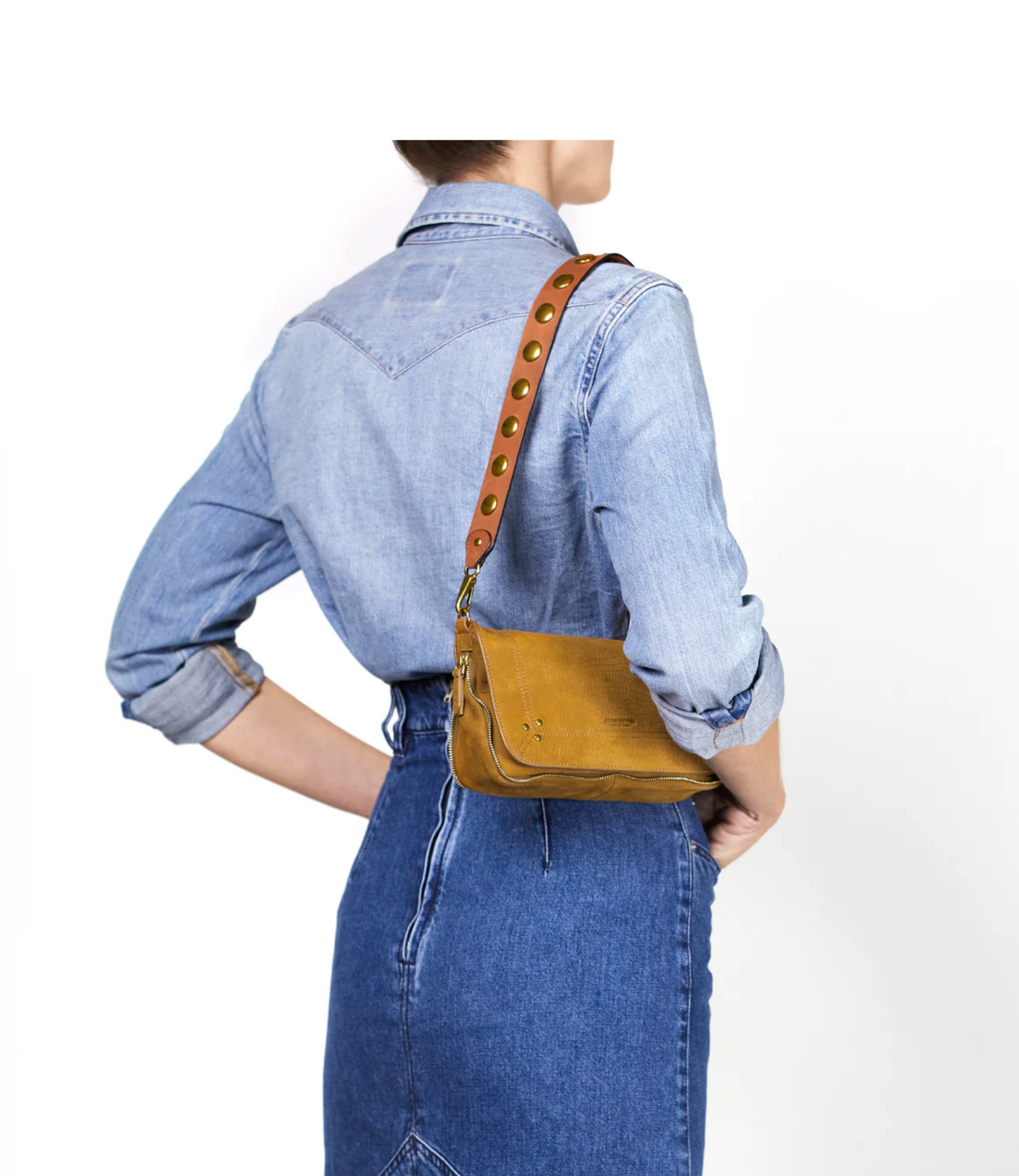 A person wears a light blue denim shirt and high-waisted skirt, carrying the Jerome Dreyfuss Anse Festival Strap—a small mustard yellow shoulder bag in genuine vegetable cowhide with a 64 cm gold-grommet leather strap—against a plain white background.