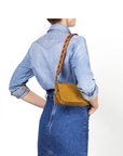 A person wears a light blue denim shirt and high-waisted skirt, carrying the Jerome Dreyfuss Anse Festival Strap—a small mustard yellow shoulder bag in genuine vegetable cowhide with a 64 cm gold-grommet leather strap—against a plain white background.