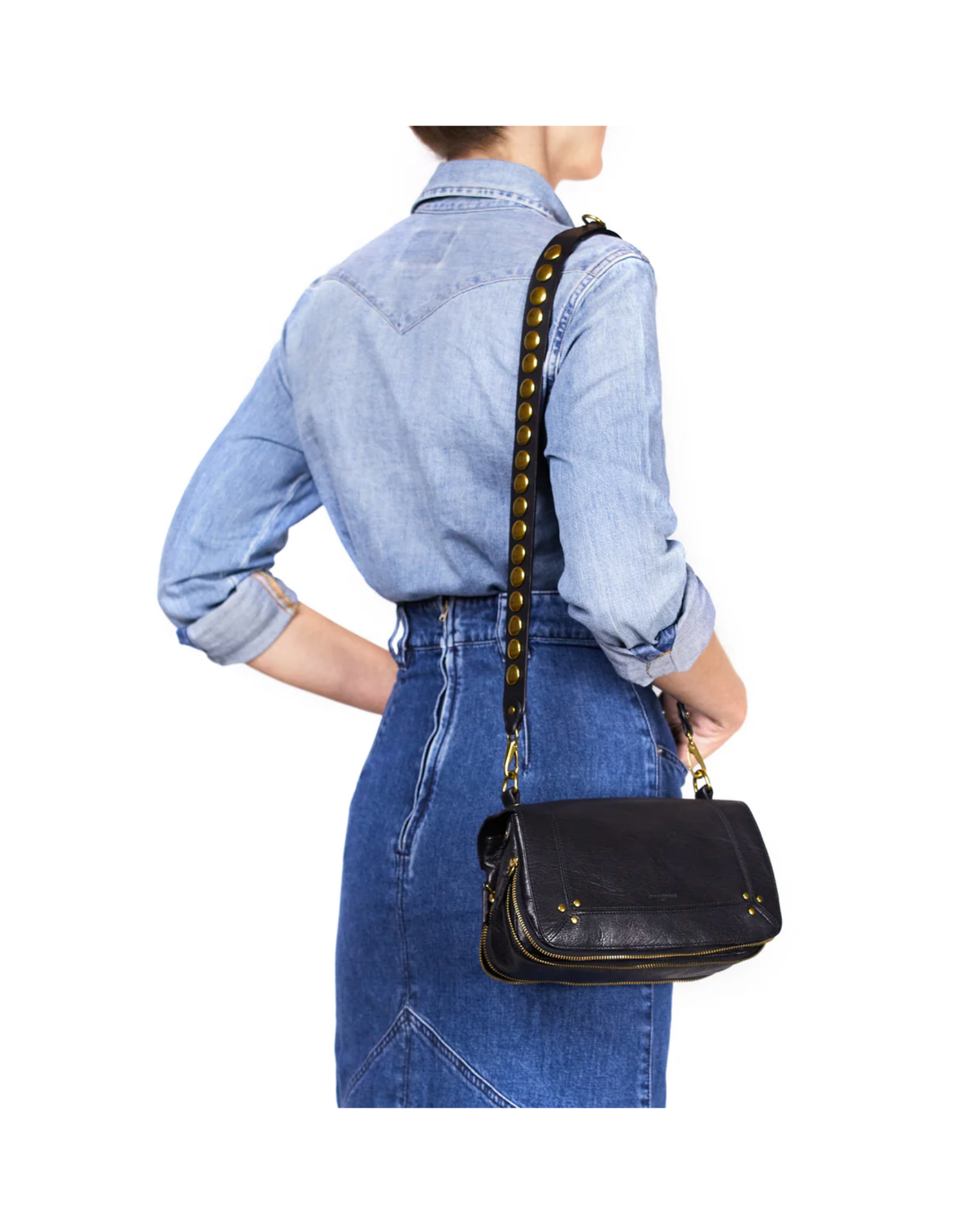 A woman in a light denim shirt and high-waisted skirt stands with her back to the camera, holding the Jerome Dreyfuss Bandouliere Festival Strap, a black leather shoulder bag with metal studs and removable shoulder strap.
