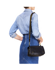 A woman in a light denim shirt and high-waisted skirt stands with her back to the camera, holding the Jerome Dreyfuss Bandouliere Festival Strap, a black leather shoulder bag with metal studs and removable shoulder strap.