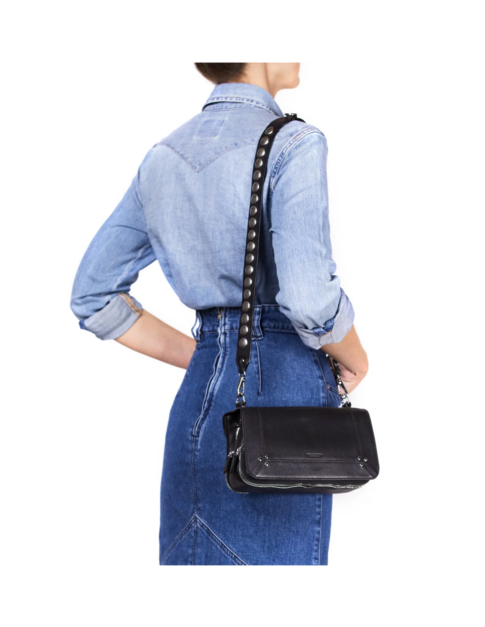 A woman in a light blue denim shirt and skirt stands with her back to the camera, carrying the Jerome Dreyfuss Bandouliere Festival Strap, a black leather shoulder bag with metal studs and a removable strap.