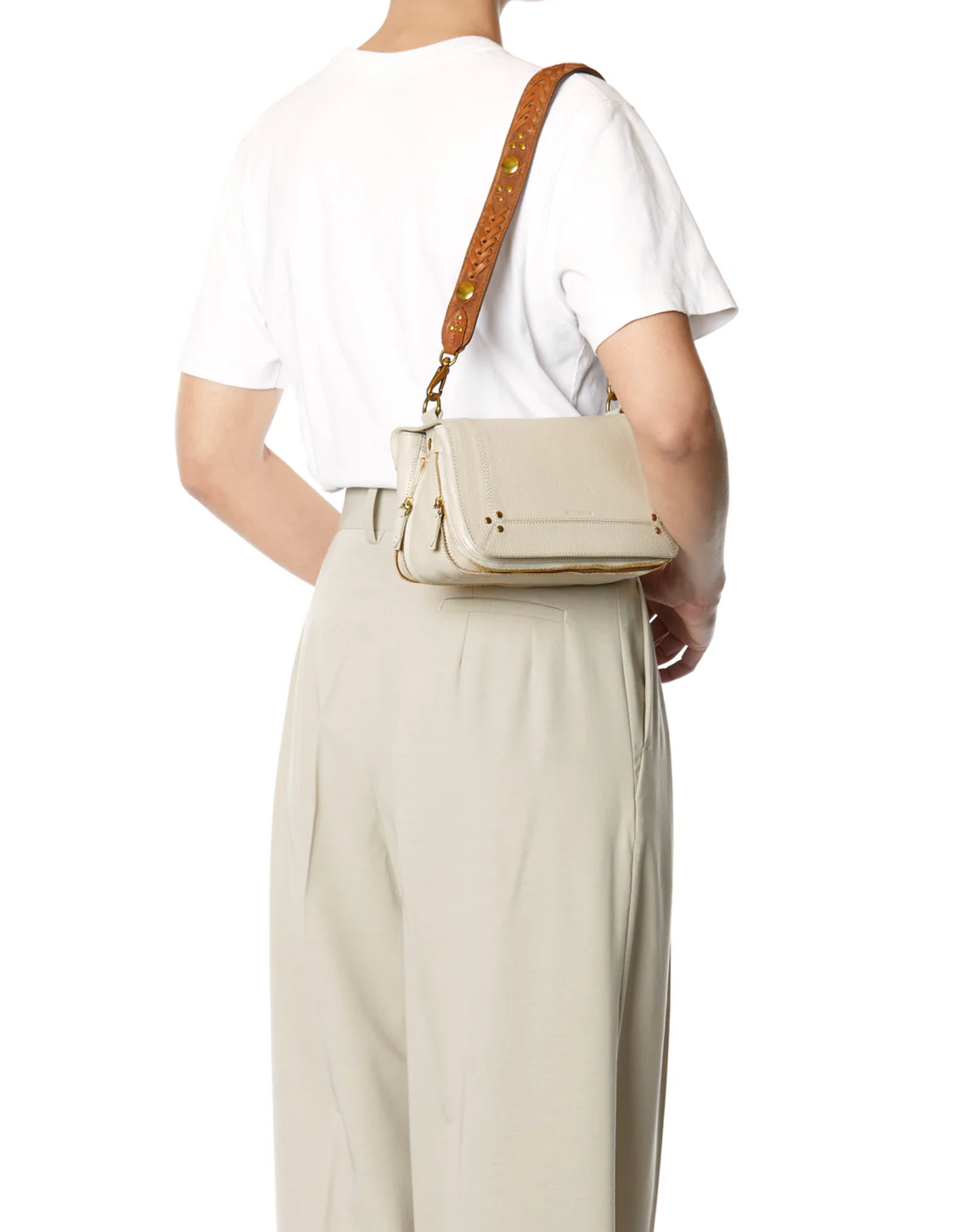 A person is seen from the back wearing a white t-shirt, high-waisted beige pants featuring the Gaucho Ceinture/Belt by Jerome Dreyfuss, and carrying a light beige crossbody bag with a wide brown strap.