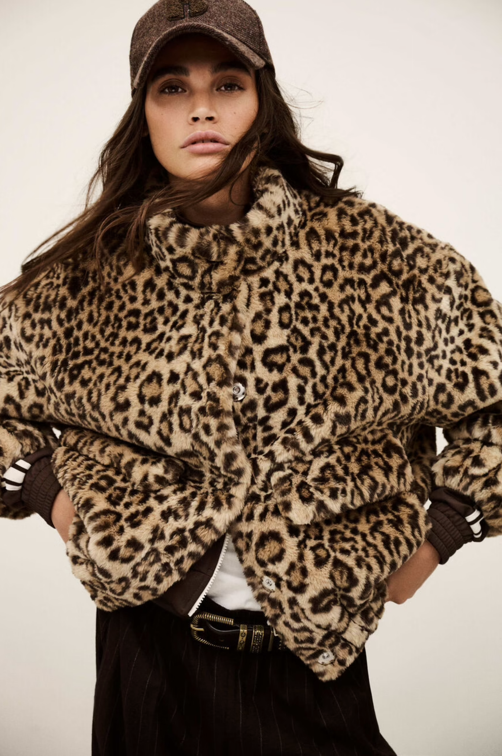 A woman models the BA&SH Coat Jaros Leopard, a brown leopard print faux fur jacket, paired with a brown cap, white top, and black pinstripe pants with a belt against a neutral background.