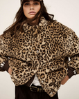 A woman models the BA&SH Coat Jaros Leopard, a brown leopard print faux fur jacket, paired with a brown cap, white top, and black pinstripe pants with a belt against a neutral background.