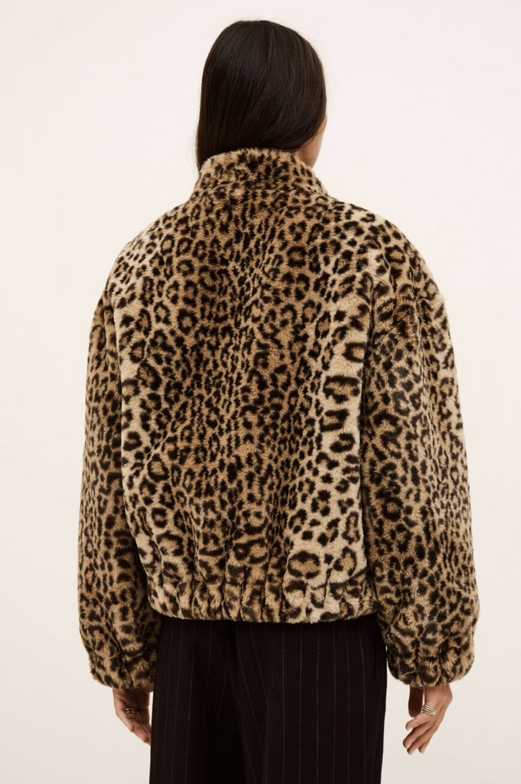 A person with long dark hair, seen from behind, wears the BA&SH Coat Jaros Leopard faux fur jacket and black pinstripe pants against a plain light background.