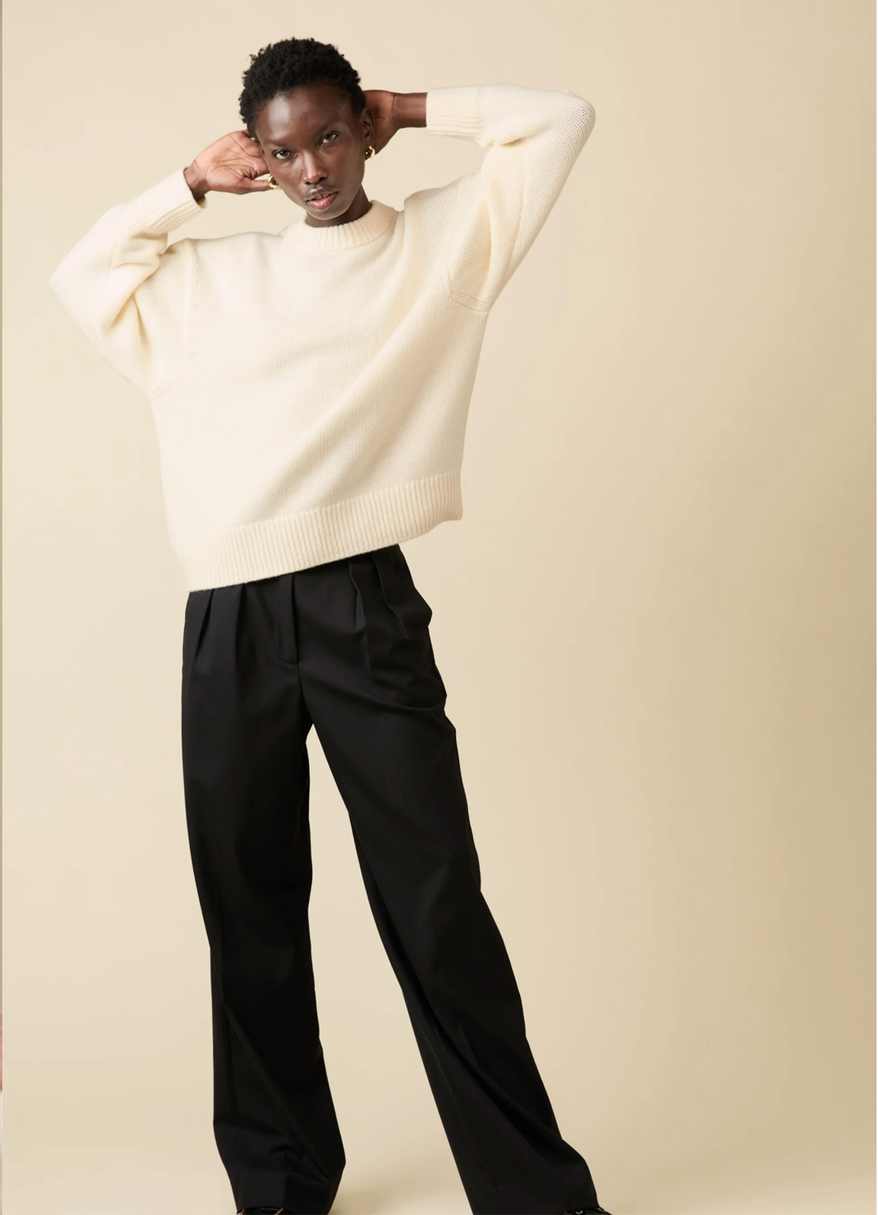 A person poses with arms raised against a beige backdrop, wearing Sold Out NYC’s Cashmere Signature Crew in cream and wide-leg black trousers.