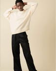 A person poses with arms raised against a beige backdrop, wearing Sold Out NYC’s Cashmere Signature Crew in cream and wide-leg black trousers.