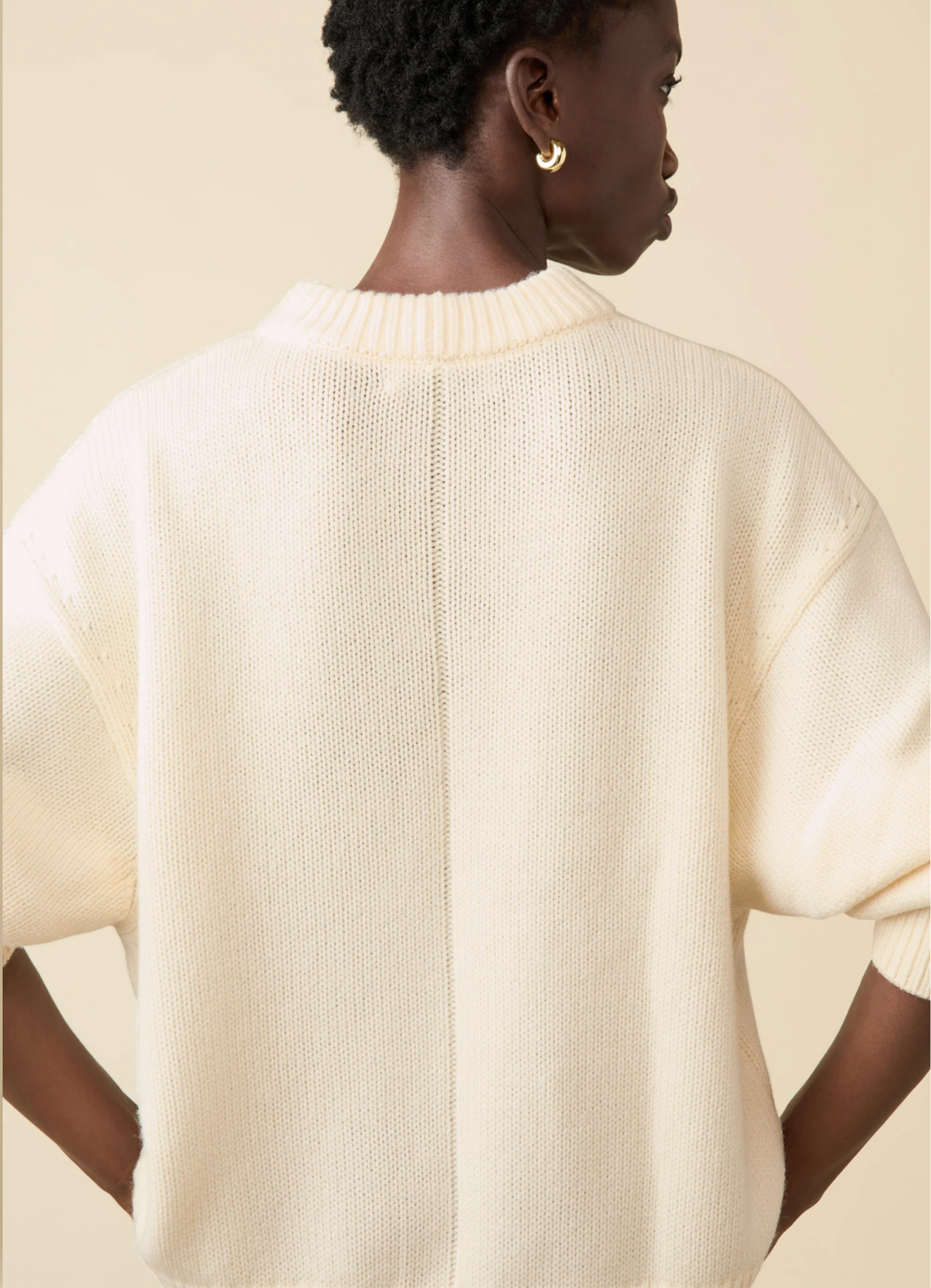 A person with short curly hair and gold hoop earrings models the Sold Out NYC Cashmere Signature Crew, an oversized cream knit sweater, shown from the back against a beige background.