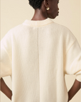 A person with short curly hair and gold hoop earrings models the Sold Out NYC Cashmere Signature Crew, an oversized cream knit sweater, shown from the back against a beige background.