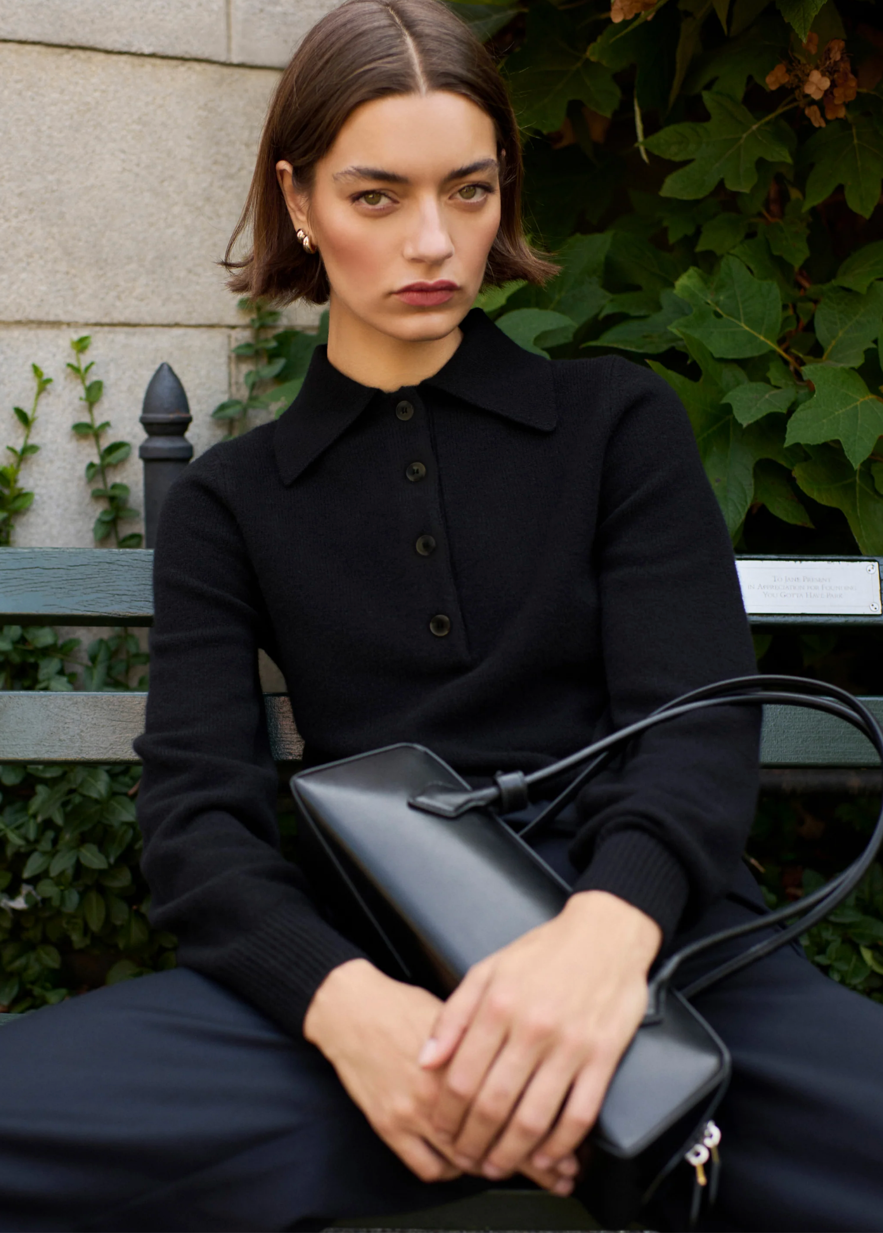 A person with short brown hair sits on a bench, wearing the Sold Out NYC Cashmere Polo and dark pants, holding a black handbag on their lap. Lush green foliage and a stone wall form the background.