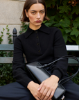 A person with short brown hair sits on a bench, wearing the Sold Out NYC Cashmere Polo and dark pants, holding a black handbag on their lap. Lush green foliage and a stone wall form the background.