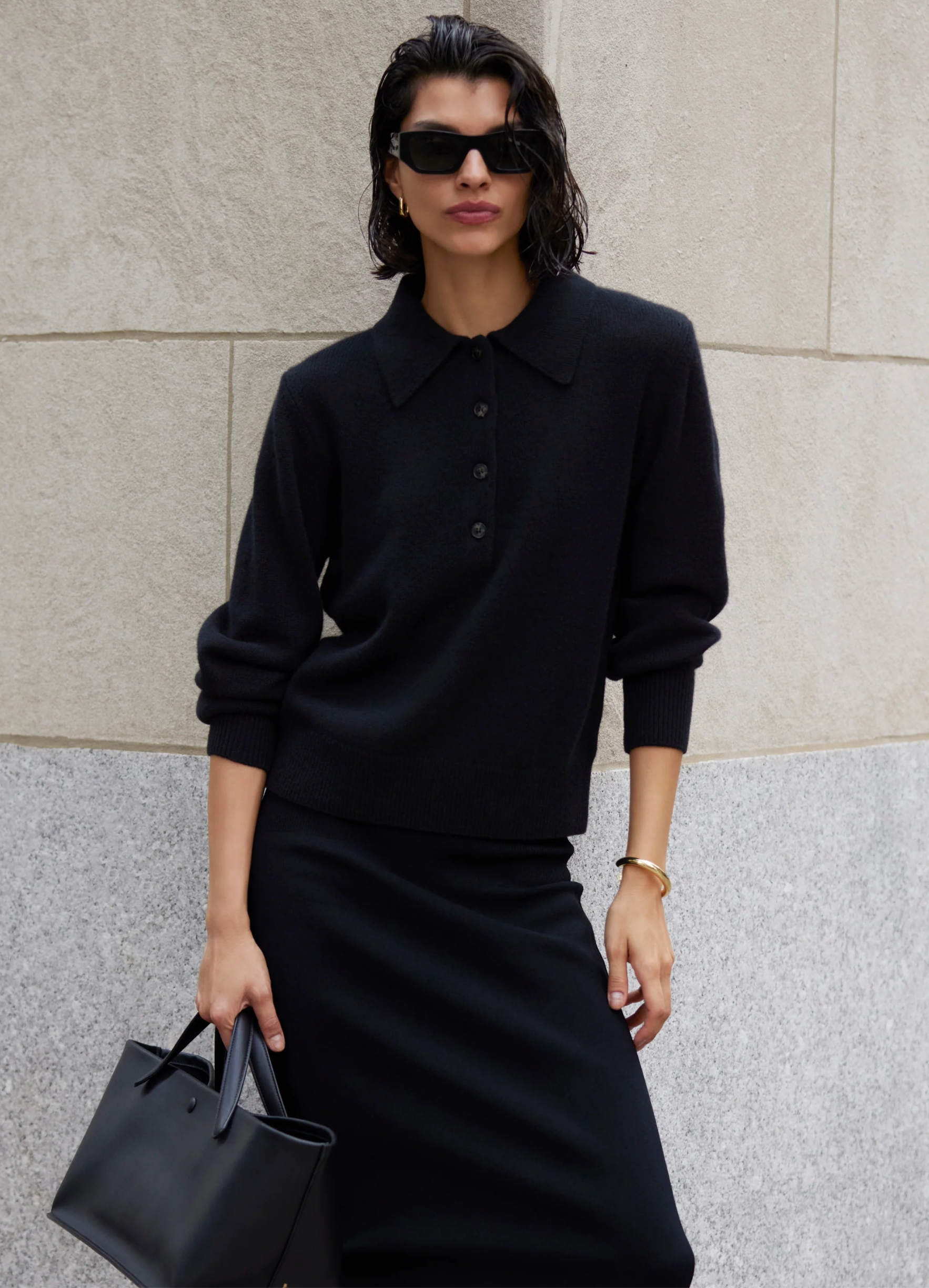 A woman in a Sold Out NYC Cashmere Polo and skirt, with black sunglasses, gold jewelry, and a black handbag stands against a stone wall.