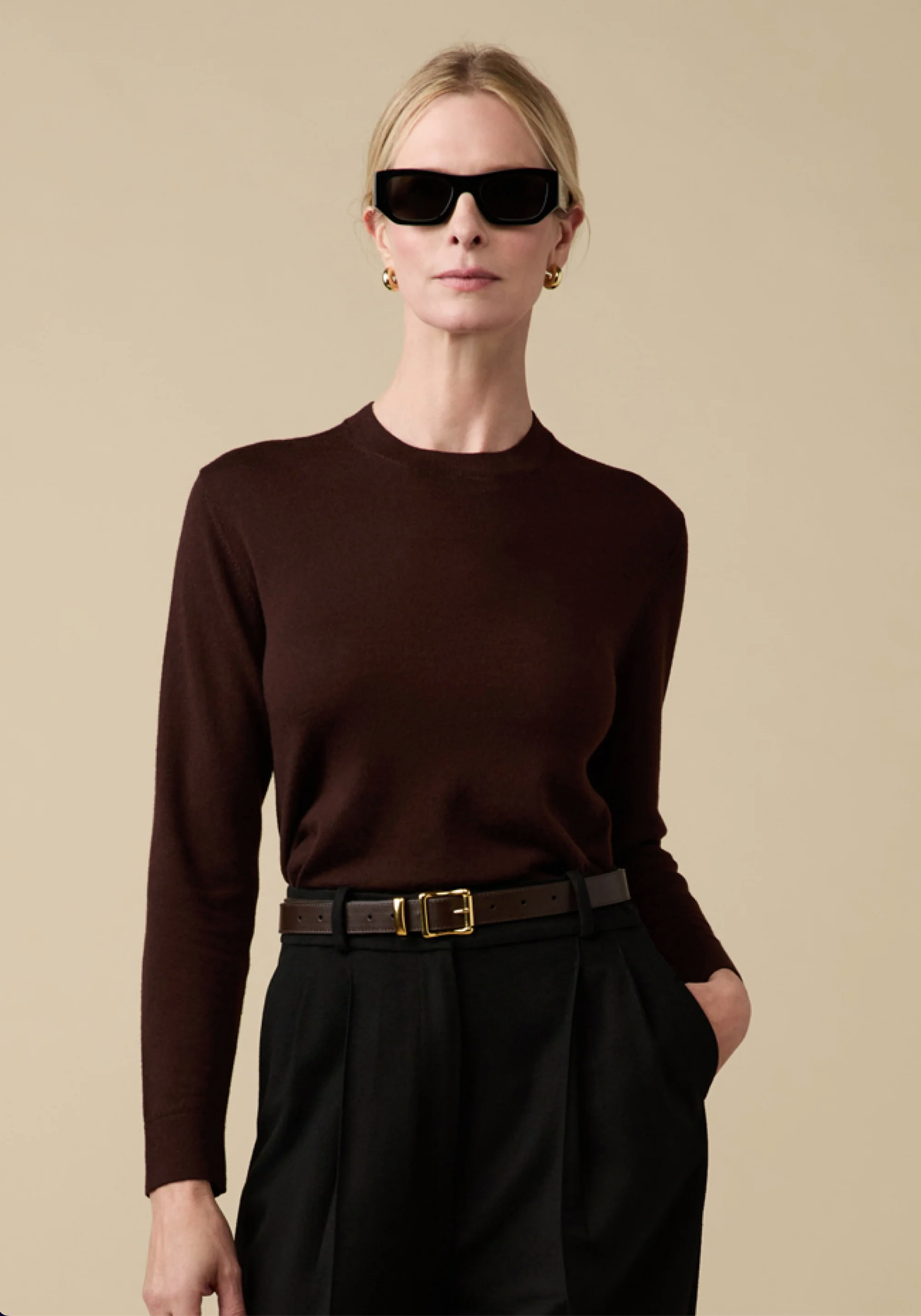 A blonde woman stands against a beige background wearing black sunglasses, gold hoop earrings, a dark brown Sold Out NYC Refined Crew sweater, and black high-waisted pants with a brown belt.