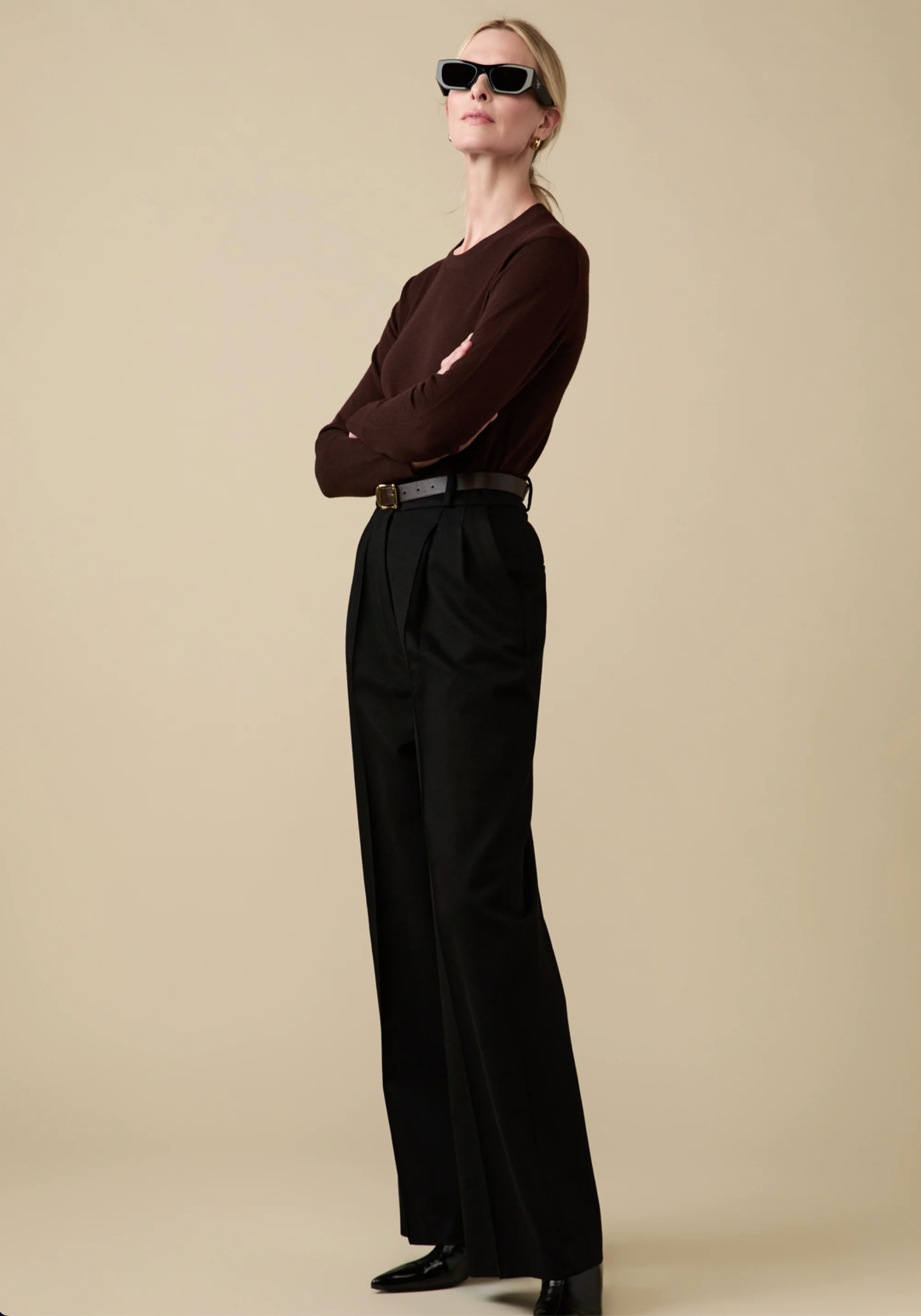 A woman confidently stands against a beige background wearing Sold Out NYC's Refined Crew sweater in dark brown, paired with high-waisted black wide-leg trousers, a belt, black shoes, and black sunglasses.