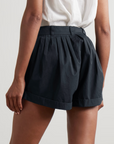 A person shown from the back wears a white top and DOEN's Paige Short—high-rise, black tailored shorts—with one hand resting by their side.