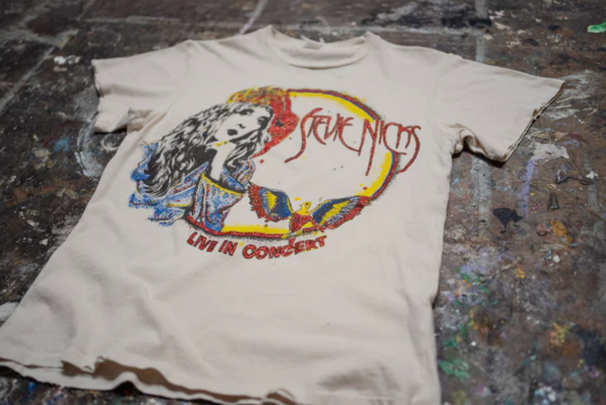 The Made Worn Stevie Nicks Tee is a cream vintage unisex 90s-fit shirt featuring a graphic of a woman, colorful designs, and "Stevie Nicks" and "Live in Concert" text on the front, shown on a paint-splattered surface.