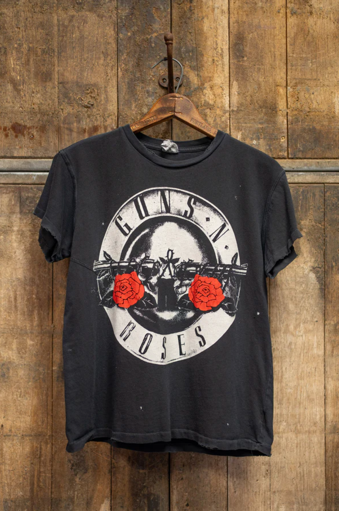 The Made Worn Guns N Roses Crew Tee, featuring two guns, two red roses, and the band’s name in a circular design, hangs on a wooden hanger against a rustic wood background—ideal for any Welcome to The Jungle fan.