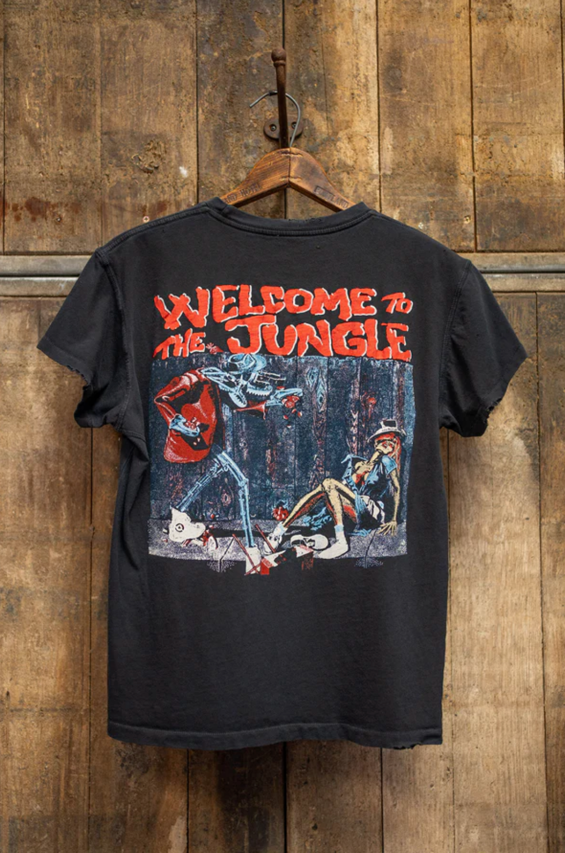A black Made Worn Guns N Roses Crew Tee hangs on a wooden wall. The back showcases bold red "WELCOME TO THE JUNGLE" text above a graphic of a skeleton attacking a man in a forest.