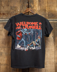 A black Made Worn Guns N Roses Crew Tee hangs on a wooden wall. The back showcases bold red "WELCOME TO THE JUNGLE" text above a graphic of a skeleton attacking a man in a forest.